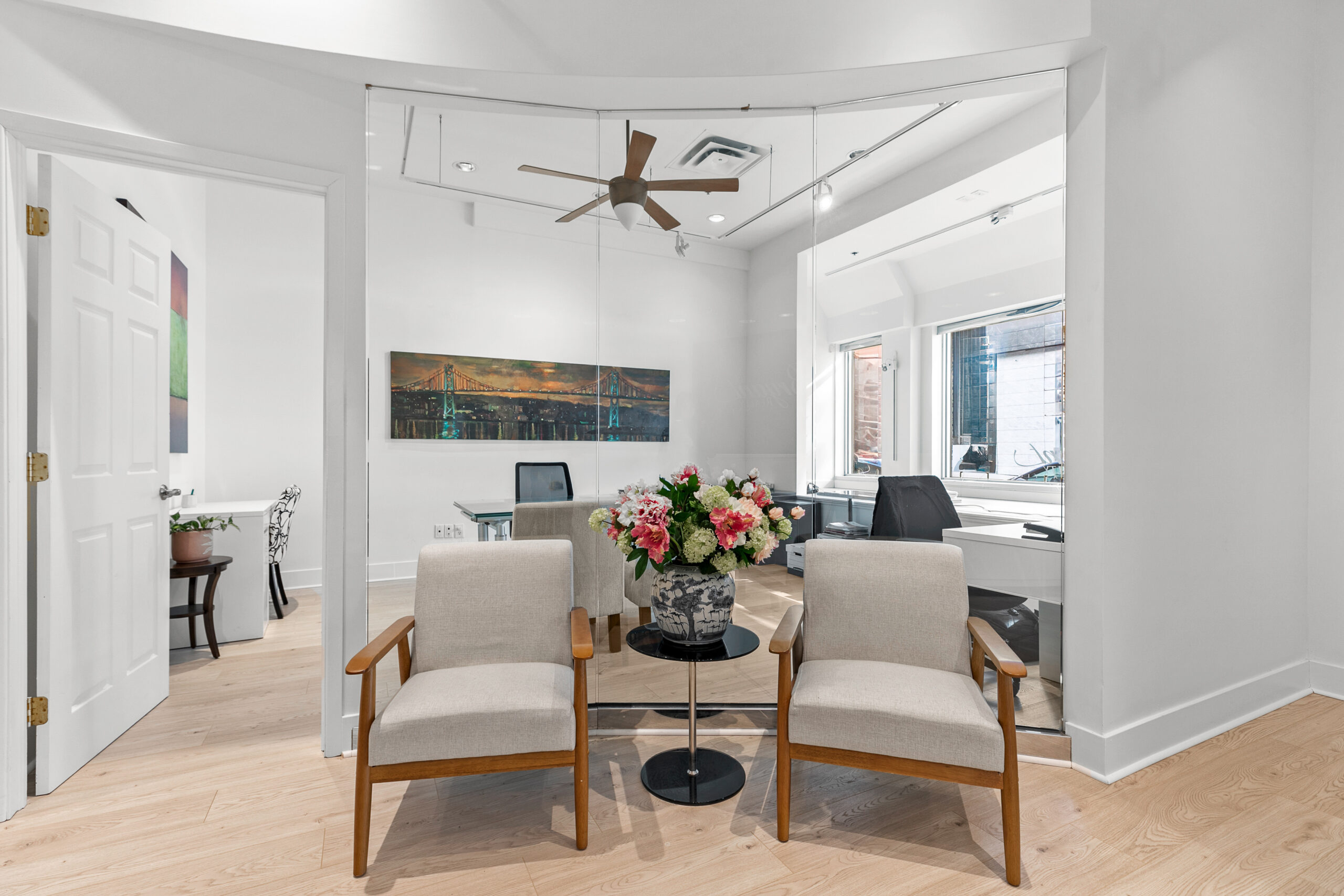 A modern office space with two beige chairs and a small round table holding a bouquet of flowers. In the background, there is a glass partition, a desk, and a large window. The room features light wood flooring and a ceiling fan.