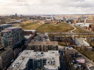 Aerial view of a cityscape with a mix of residential and commercial buildings in the foreground. A large open green space, roads, and additional buildings are visible in the background under a cloudy sky.