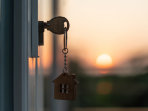 A key with a small house-shaped keychain is inserted in a door lock, with a blurred sunset in the background. The warm colors of the sunset create a serene atmosphere.