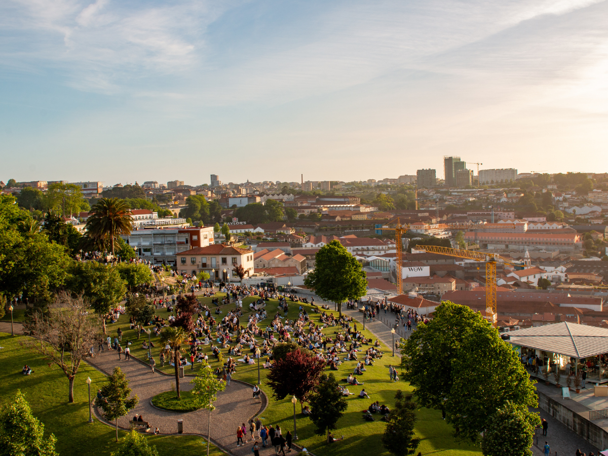 Aerial view of a park crowded with people sitting on the grass, surrounded by trees and city buildings under a clear blue sky. The scene captures a lively urban space during daylight hours.