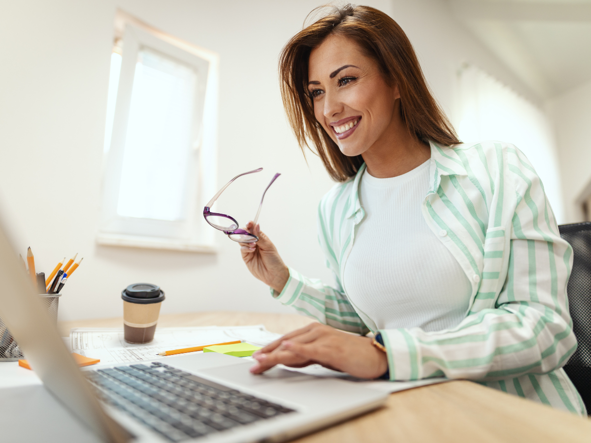 Woman smiling while working on a laptop at a desk. She's holding glasses in one hand and the desk has a coffee cup, pens, and papers. She's wearing a striped shirt and seated in a room with a window in the background.