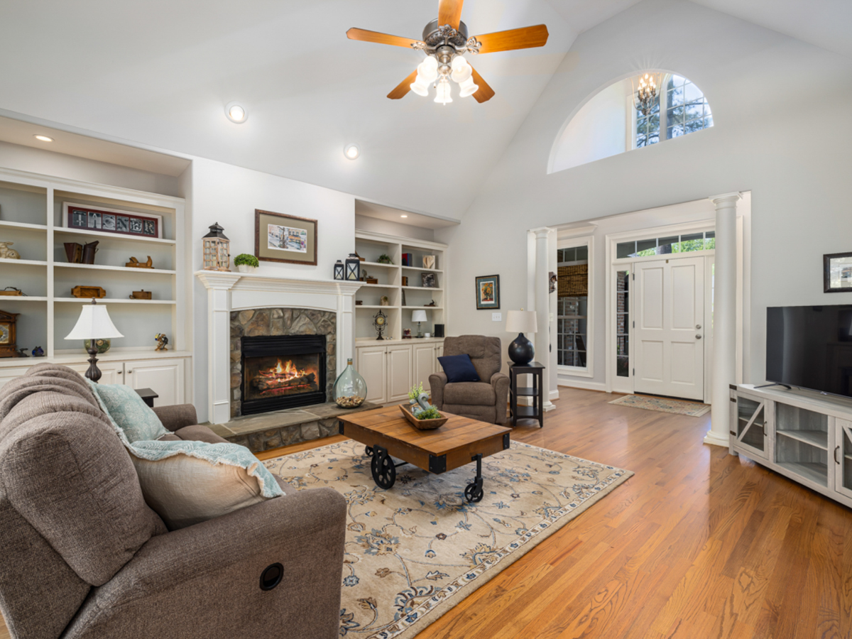 Spacious living room with vaulted ceiling, featuring a stone fireplace, two brown sofas, a wooden coffee table, built-in shelves, and a flat-screen TV. Large windows provide natural light, and a ceiling fan is above. Hardwood floors and a patterned rug add warmth.