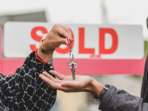A person hands over a key with a house-shaped keychain to another person's open hand. A "SOLD" sign is visible in the background, partially blurred. The person receiving the key is wearing a dark jacket.