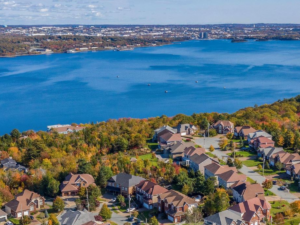 Aerial view of a suburban neighborhood with houses surrounded by autumn trees, overlooking a large blue lake with a distant city and sky in the background.