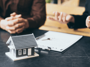 Two people sit at a table with a contract and keys; a small model house is in the foreground, suggesting a real estate or home buying discussion.
